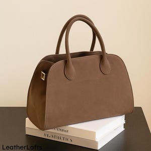 May include: A brown handbag with two rounded handles and a rectangular shape. The bag has a gold-coloured clasp and is sitting on top of a stack of books. The bag is made of a suede-like material.
