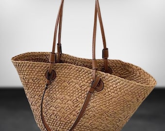 May include: A woven straw tote bag with brown leather handles and accents. The bag has a wide opening and a textured weave pattern. The handles are attached with leather loops and stitching. The bag is a natural tan color.
