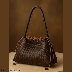 May include: A dark brown woven leather handbag with a braided handle and drawstring closure. The interior is a lighter brown fabric. The bag has a rounded shape and is set against a neutral background. The brand name "LeatherLofts" is visible at the bottom.