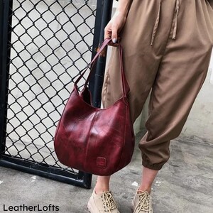 May include: A burgundy leather shoulder bag with a textured finish. The bag has a curved shape and a single shoulder strap. The bag is held by a person wearing tan pants and shoes. The words "LeatherLofts" are at the bottom left.