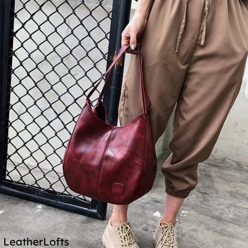 May include: A burgundy leather shoulder bag with a textured finish. The bag has a curved shape and a single shoulder strap. The bag is held by a person wearing tan pants and shoes. The words "LeatherLofts" are at the bottom left.