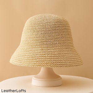 Puede incluir: Un sombrero de cubo beige tejido con ala ancha. El sombrero se exhibe sobre un pedestal blanco, sobre un fondo neutro. La textura y la forma del sombrero son claramente visibles, lo que sugiere un accesorio de verano.