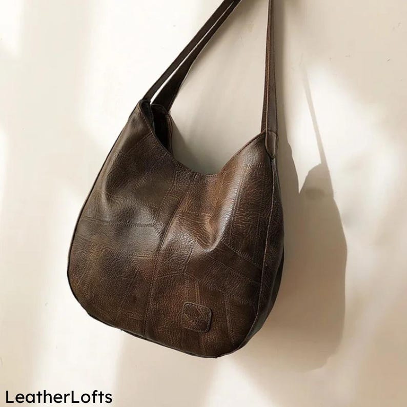 May include: A brown leather shoulder bag with a patchwork design. The bag has a curved shape and long straps. The text "LeatherLofts" is visible at the bottom left of the image.