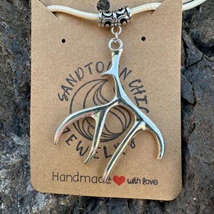 Könnte beinhalten: Halskette mit silberfarbenem Geweih-Anhänger an einer cremefarbenen Kordel. Der Anhänger hängt an einer braunen Karte mit dem Text "SANDTOWN CHIC JEWELRY Handmade with love". Die Halskette wird auf einer strukturierten, dunkelbraunen Oberfläche präsentiert.