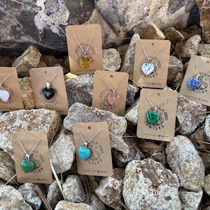 May include: A selection of heart-shaped gemstone pendants on silver chains, presented on individual kraft paper cards. The gemstones are in various colours, including white, black, yellow, pink, green, blue, and turquoise. The cards have the text "Sandtown Chic Jewelry".