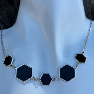 May include: A silver-toned necklace featuring a series of black, hexagon-shaped pendants. The geometric design is set against a white background, highlighting the contrast between the dark pendants and the bright metal chain.