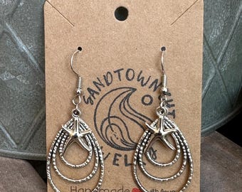 May include: Silver-toned dangle earrings featuring a starfish design. Each earring has three nested teardrop-shaped hoops and a starfish charm at the top. The earrings are displayed on a brown card with the text "SANDTOWN JEWEL" and "Handmade with love."