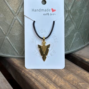 May include: Gold-colored arrowhead pendant necklace on a black cord. The pendant is attached to a white card that reads "Handmade with love." The necklace is displayed on a wooden surface.