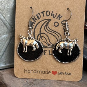 May include: Silver-tone wolf earrings with black felt backing. The earrings hang from silver hooks and are displayed on a brown card with the words "Handmade with love."