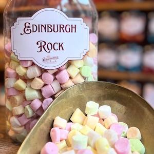 Könnte beinhalten: Ein klares Glas mit bunten Edinburgh Rock-Bonbons, versehen mit einem weißen und weinroten Etikett. Eine kleine Messingschaufel enthält einen Teil der pastellfarbenen Süßigkeiten. Der Behälter hat einen schwarzen Deckel.