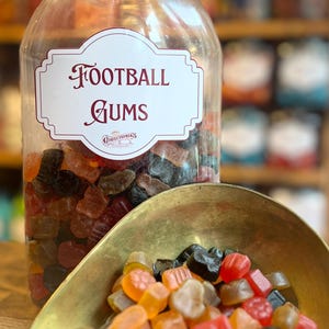 Pode incluir: Um frasco de plástico transparente cheio de gomas coloridas em forma de futebol americano. O frasco tem uma tampa preta e um rótulo que diz "Football Gums". Uma pequena concha de latão contém uma pilha de doces.