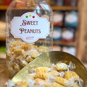 Könnte beinhalten: Ein klares Glas mit schwarzem Deckel mit der Aufschrift "Sweet Peanuts", gefüllt mit einzeln verpackten Süßigkeiten. Eine Messingkelle enthält weitere Erdnussbonbons. Regale mit Gläsern sind im Hintergrund.