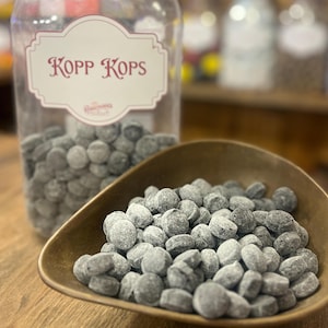 May include: A clear glass jar with a black lid and a label that reads "Kopp Kops" is filled with gray candies. A small, bronze-colored bowl holds a pile of the same candies. The candies are small, round, and appear to be dusted.