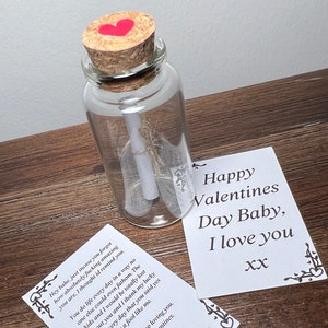 Könnte beinhalten: Eine kleine Glasflasche mit einem Korkverschluss und einem roten Herzen auf dem Korken. Die Flasche enthält ein zusammengerolltes Stück Papier. Eine Karte mit dem Text "Happy Valentines Day Baby, I love you xx" liegt neben der Flasche.