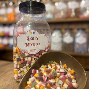 Pode incluir: Um frasco grande e transparente de doces Dolly Mixture com tampa preta e rótulo. Os doces são uma mistura de pedaços coloridos de vários tamanhos e cores. Uma pequena tigela marrom contém uma porção dos doces.