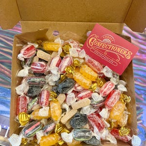 May include: A selection of wrapped sweets in a cardboard box. The sweets are in a variety of colours, including red, yellow, black, and white. A red card with the text "Coque Confectioners Northumberland" is visible.