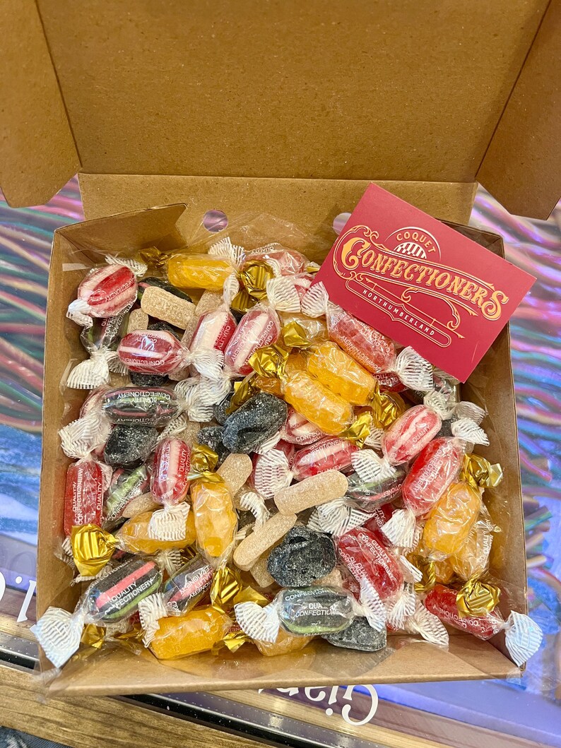 May include: A selection of wrapped sweets in a cardboard box. The sweets are in a variety of colours, including red, yellow, black, and white. A red card with the text "Coque Confectioners Northumberland" is visible.