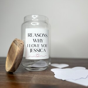 May include: A clear glass jar with a wooden lid. The jar has a white label that says "Reasons Why I Love You Jessica" with a small red heart. There are white heart-shaped pieces of paper next to the jar.