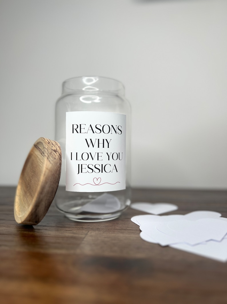 May include: A clear glass jar with a wooden lid. The jar has a white label that says "Reasons Why I Love You Jessica" with a small red heart. There are white heart-shaped pieces of paper next to the jar.