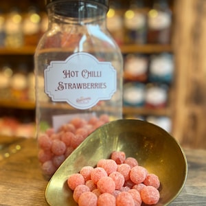 Op de afbeelding: Een doorzichtige glazen pot met een zwarte deksel en een label met de tekst "Hot Chilli Strawberries" is gevuld met roze snoepjes. Een messing schep bevat een stapel van dezelfde snoepjes. Schappen met potten staan op de achtergrond.