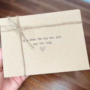 May include: A brown paper envelope with twine tied around it. The envelope has the text "open when the day was just way too long" printed on it with a small heart symbol.