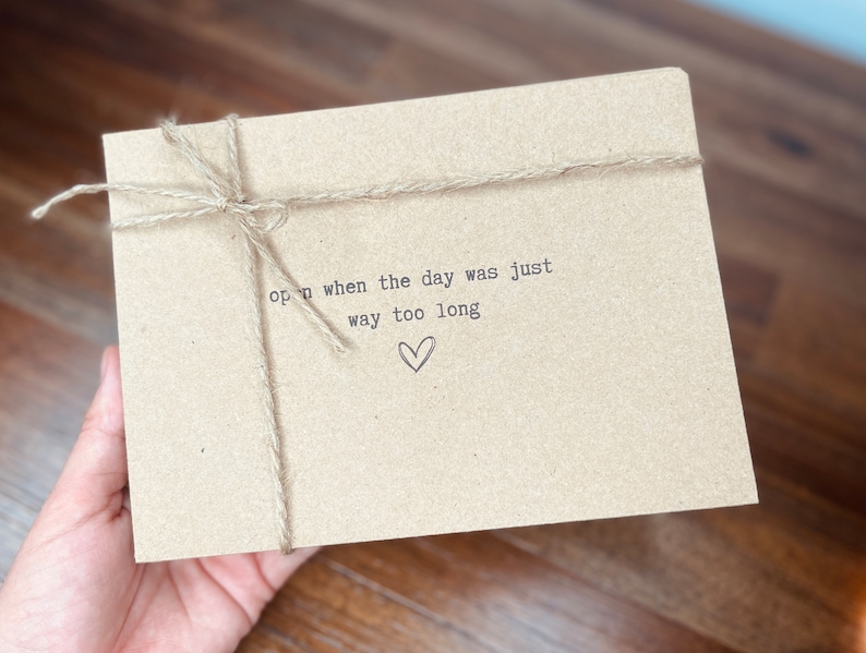 May include: A brown paper envelope with twine tied around it. The envelope has the text "open when the day was just way too long" printed on it with a small heart symbol.