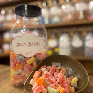 May include: A clear glass jar with a black lid labeled "Jelly Babies" sits next to a small, bronze-colored scoop filled with colorful candies. The candies are in various colors, including red, orange, yellow, green, and gray. The background shows shelves of jars.