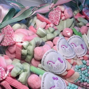 May include: A collection of clear cellophane bags filled with colorful candies. The candies are primarily pink, white, and green, with a variety of shapes and textures. Each bag is tied with pink and green ribbons. Round stickers with a unicorn and text are visible.