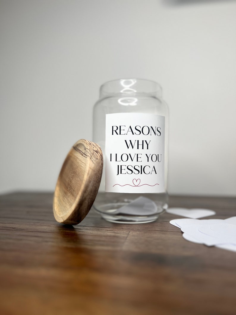 May include: A clear glass jar with a white label that reads "Reasons Why I Love You Jessica" and a small red heart. The jar is open and there are white paper hearts on a wooden surface.