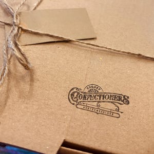 May include: A brown cardboard box tied with twine and a blank tag. The box features the printed logo of "Coquet Confectioners Northumberland". The box is closed and ready for gifting, suggesting a food item.