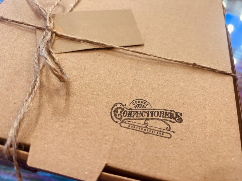 May include: A brown cardboard box tied with twine and a blank tag. The box features the printed logo of "Coquet Confectioners Northumberland". The box is closed and ready for gifting, suggesting a food item.