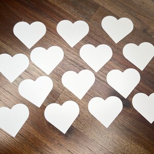 May include: A collection of 14 white paper hearts arranged in a pattern on a wood surface.
