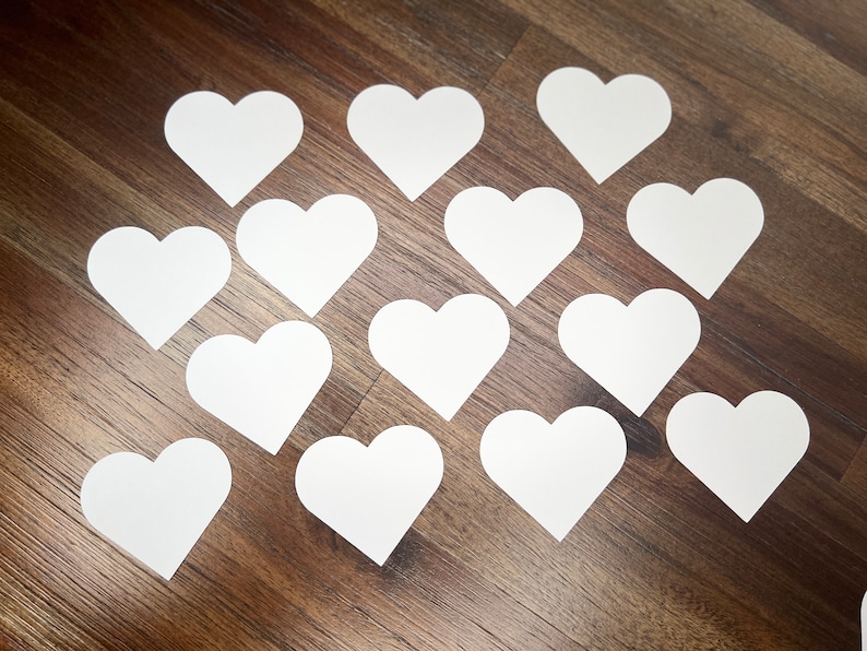 May include: A collection of 14 white paper hearts arranged in a pattern on a wood surface.