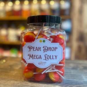 May include: A jar of pear drop lollipops. The clear jar has a black lid and a label that reads "Pear Drop Mega Lolly". The lollipops are red and yellow, and are on white sticks. The background is blurred, suggesting a shop setting.