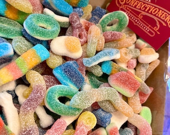 May include: A box filled with a variety of colorful, sugared gummy candies. The assortment includes rings, worms, and other shapes in shades of green, blue, yellow, and red. A red card with the text "Confectioners" is visible.