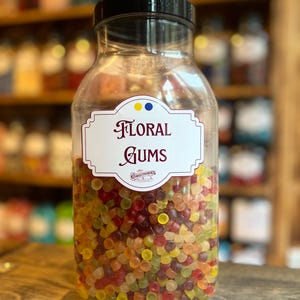 May include: A clear glass jar filled with colorful floral gums. The jar has a black lid and a white label with the words "FLORAL GUMS" in elegant script. The gums are a mix of red, yellow, green, and orange.