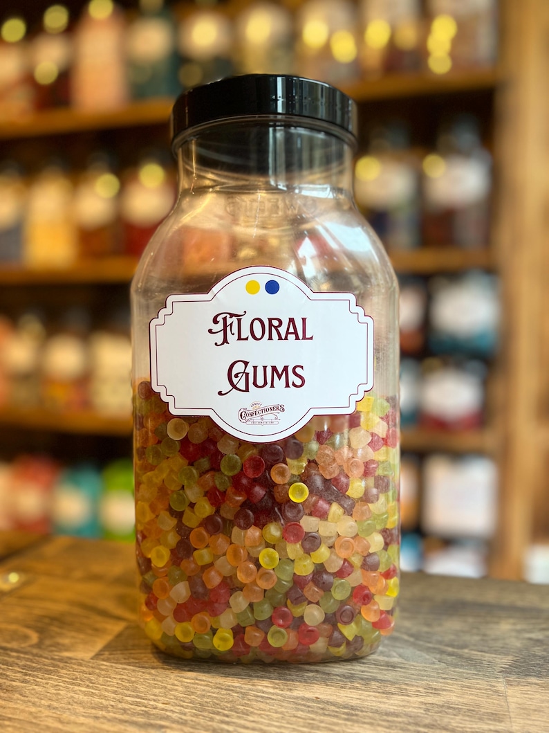 May include: A clear glass jar filled with colorful floral gums. The jar has a black lid and a white label with the words "FLORAL GUMS" in elegant script. The gums are a mix of red, yellow, green, and orange.