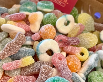 May include: A box filled with a colorful assortment of sugared gummy candies. The candies are in various shapes, including rings, worms, and fruit slices, and in colors like green, orange, yellow, and pink. A red card with the text "Confectioners" is in the box.