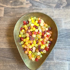 May include: A brass-colored, teardrop-shaped bowl filled with colorful, square-shaped candies. The candies are a mix of red, yellow, orange, green, and purple. The bowl sits on a wooden surface.