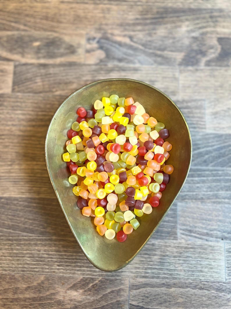 May include: A brass-colored, teardrop-shaped bowl filled with colorful, square-shaped candies. The candies are a mix of red, yellow, orange, green, and purple. The bowl sits on a wooden surface.