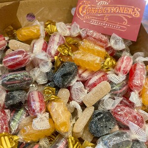 May include: A selection of wrapped sweets in a cardboard box. The candies are in various colours, including red, yellow, and black. A red card with gold lettering that reads "Confectioners" is visible. The sweets are in clear and gold wrappers.
