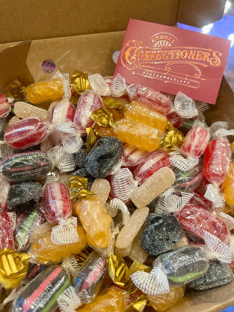 May include: A selection of wrapped sweets in a cardboard box. The candies are in various colours, including red, yellow, and black. A red card with gold lettering that reads "Confectioners" is visible. The sweets are in clear and gold wrappers.
