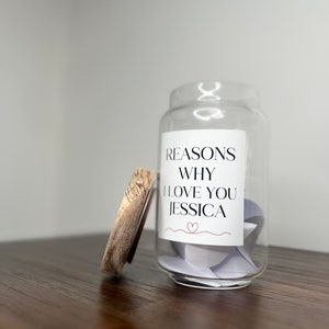 May include: A glass jar with a wooden lid filled with small pieces of paper. The jar has a label that reads "Reasons Why I Love You Jessica" with a small heart symbol.