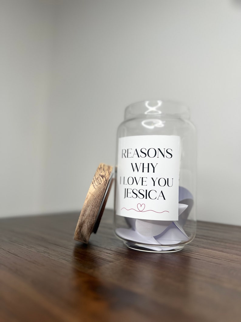 May include: A glass jar with a wooden lid filled with small pieces of paper. The jar has a label that reads "Reasons Why I Love You Jessica" with a small heart symbol.