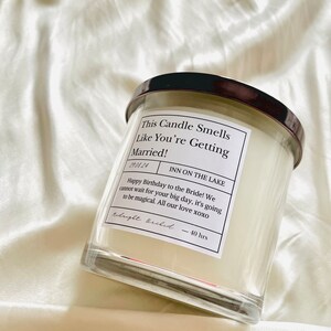 Puede incluir: Un tarro de vela de vidrio transparente con una tapa negra. La etiqueta dice "This Candle Smells Like You're Getting Married!" y "INN ON THE LAKE". La etiqueta tambi&eacute;n incluye una fecha, "19.04.24", y un mensaje deseando un feliz cumplea&ntilde;os a la novia. La vela est&aacute; etiquetada como "Midnight Orchid" y tiene un tiempo de combusti&oacute;n de 40 horas.