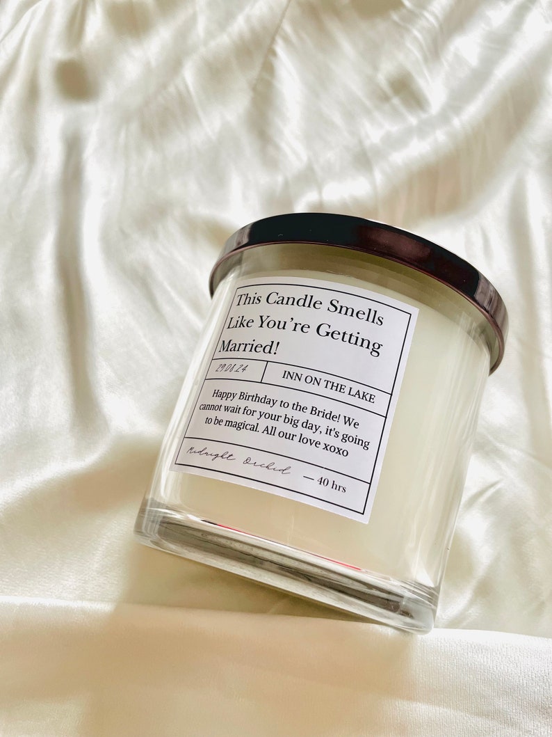 Puede incluir: Un tarro de vela de vidrio transparente con una tapa negra. La etiqueta dice "This Candle Smells Like You're Getting Married!" y "INN ON THE LAKE". La etiqueta tambi&eacute;n incluye una fecha, "19.04.24", y un mensaje deseando un feliz cumplea&ntilde;os a la novia. La vela est&aacute; etiquetada como "Midnight Orchid" y tiene un tiempo de combusti&oacute;n de 40 horas.