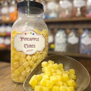 Op de afbeelding: Een heldere glazen pot gevuld met gele ananasblokjes, met het label "Pineapple Cubes". Een kleine, donkere kom bevat een stapel van dezelfde snoepjes. De achtergrond toont planken met potten gevuld met diverse snoepjes.