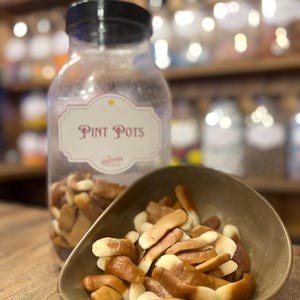 Pode incluir: Um frasco de plástico transparente com tampa preta rotulado "Pint Pots" cheio de doces castanhos e brancos. Uma pequena tigela de cor bronze contém uma porção dos doces. Os doces têm a forma de pequenos troncos castanhos e brancos.