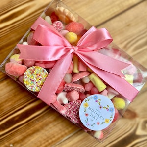 May include: A clear box filled with assorted candies, tied with a pink satin bow. The candies are in shades of pink, red, and yellow. A round sticker with the text "Happy Mother's Day" is visible.