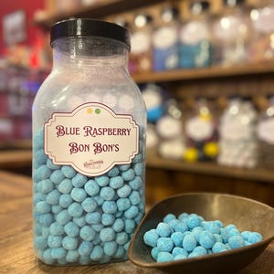Puede incluir: Un tarro de plástico transparente lleno de caramelos de frambuesa azul, con tapa negra. La etiqueta dice "Blue Raspberry Bon Bons". Un pequeño cuenco color bronce contiene una porción de los caramelos. Los caramelos son pequeños, redondos y de color azul claro.